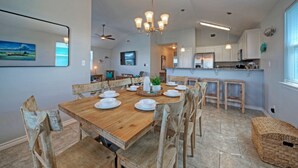 Un comedor con una mesa de madera dispuesta para seis personas, rodeada de sillas de madera. Un candelabro cuelga encima, y se observa una cocina moderna al fondo.