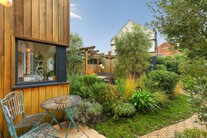 A wooden building with a window, a small patio with a metal table and chairs, and a garden with various plants and a brick pathway. - Convenient 3Bed Near Bingham Station & Town Centre (Bingham)