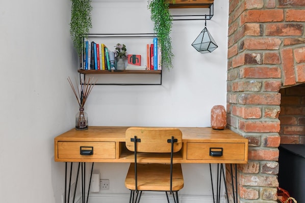 A wooden desk with two chairs, a bookshelf with books and a cactus, a hanging plant, and a brick wall.