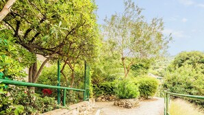 Un sendero de piedra que atraviesa un exuberante jardín con diversas árboles y arbustos, bordeado por un muro de piedra y barandillas metálicas verdes.