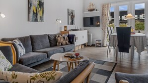 A modern living room with a grey sofa, a coffee table, and a dining area with a table and chairs. There are abstract paintings on the walls, a TV mounted above the sofa, and a ship model on a shelf. - "Benthe" - 100m to the fjord by Interhome (Vestervig)