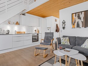 Une cuisine moderne avec des meubles blancs, un plafond en bois et un coin repas comprenant un canapé gris et une petite table blanche.