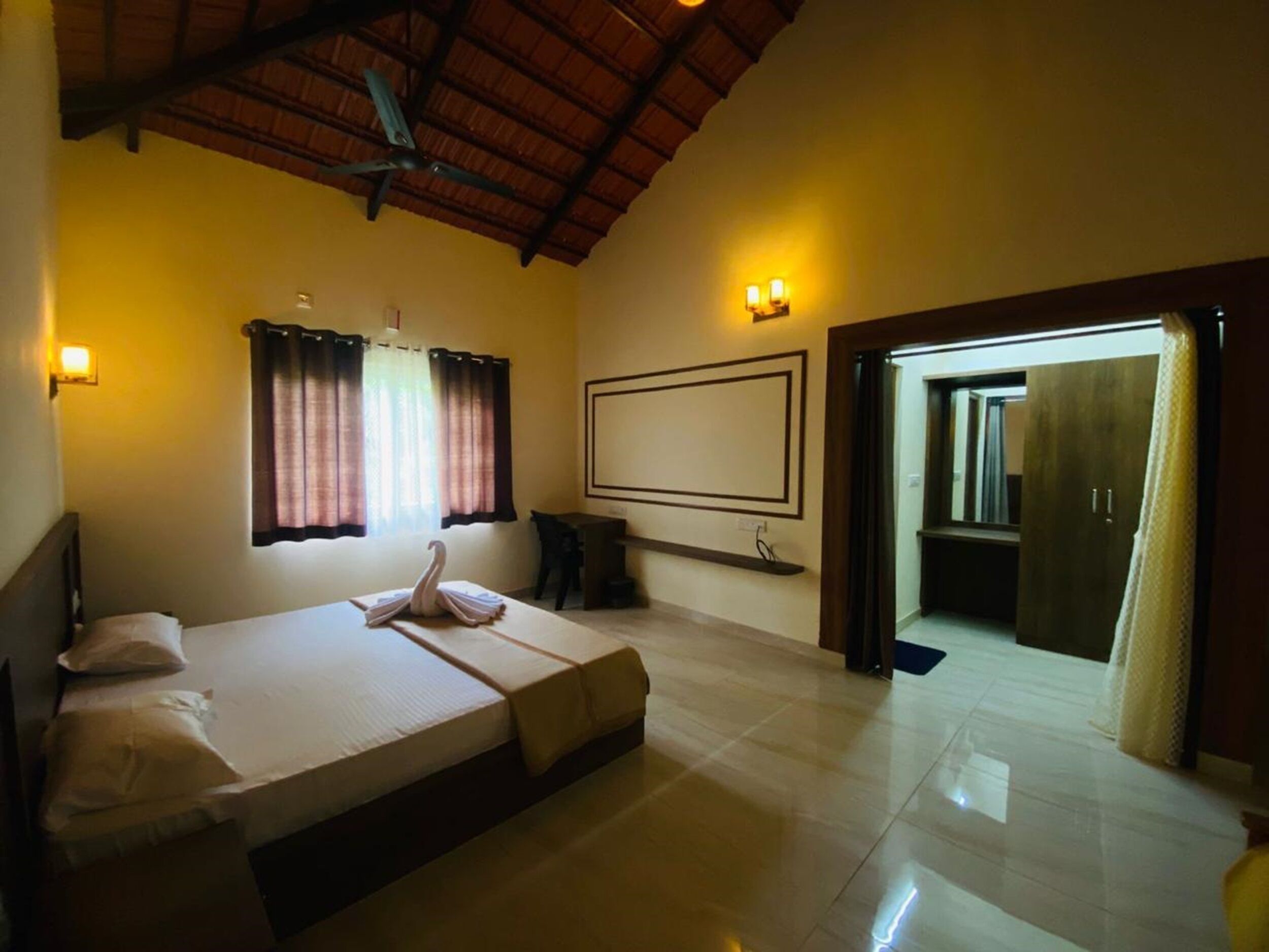 Quarto de hotel com cama, escrivaninha, cadeira, janela com cortinas, luminária na parede e porta que dá para outro cômodo.