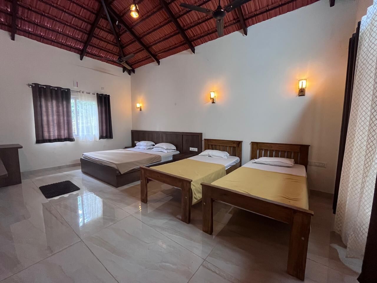 A hotel room with three single beds, a wooden bench, a desk, and a window with curtains.