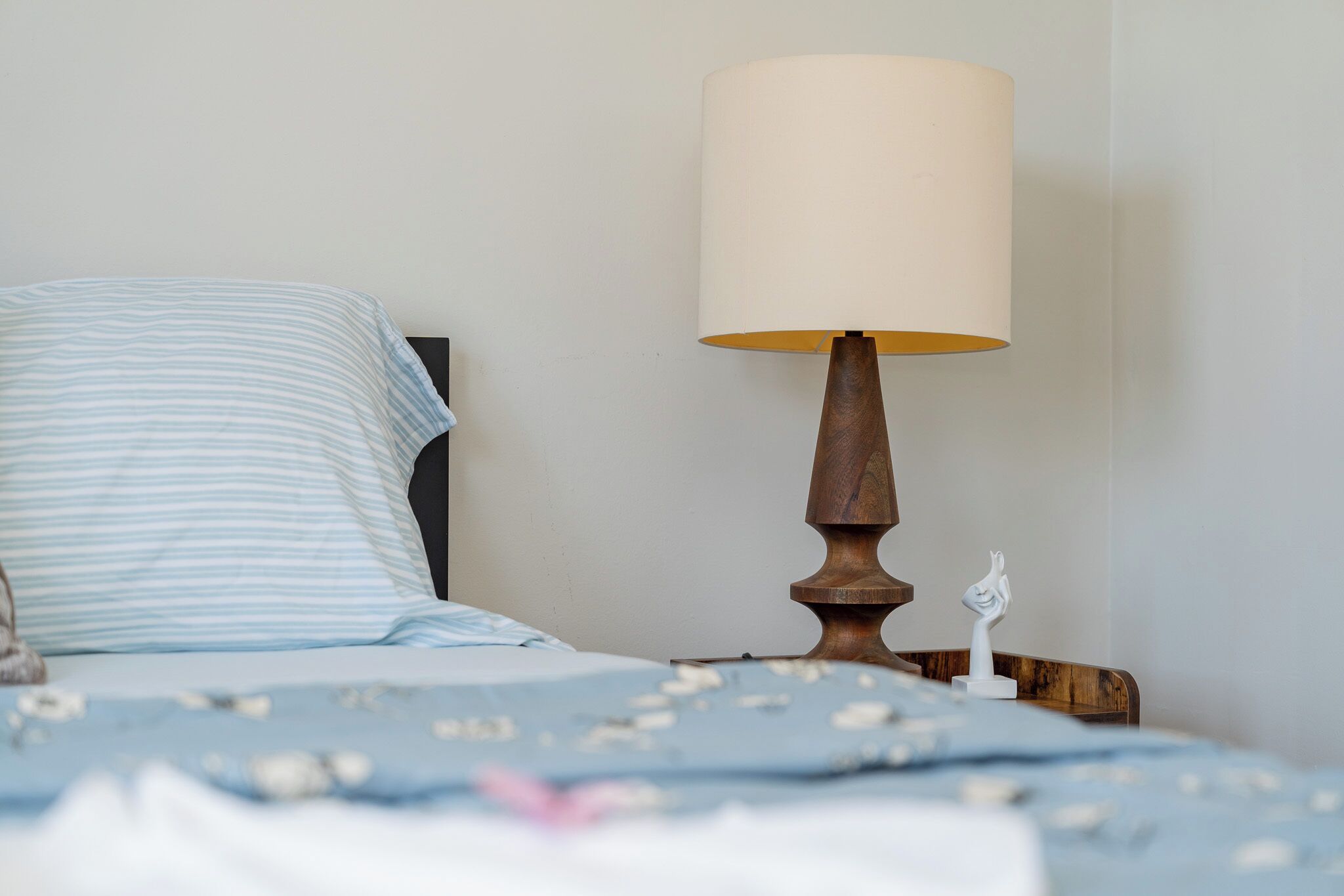 Una cama con un almohadón a rayas y una mesita de noche de madera con una lámpara blanca.