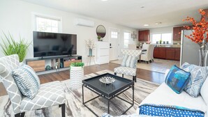 Una sala de estar moderna con un televisor de pantalla plana, un sofá y dos sillones. Hay un comedor con una cocina al fondo.