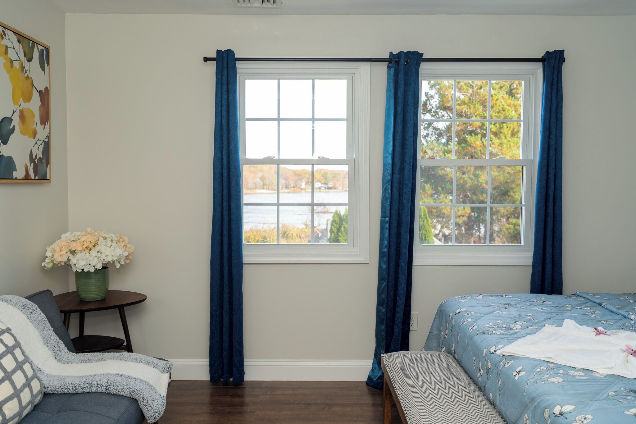 Un dormitorio con una cama, una silla, una mesita con un jarrón de flores y una ventana con cortinas.