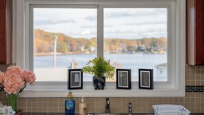 Una cocina con vista al lago y follaje otoñal a través de la ventana, una planta en maceta y dos frases motivacionales enmarcadas en el alféizar.
