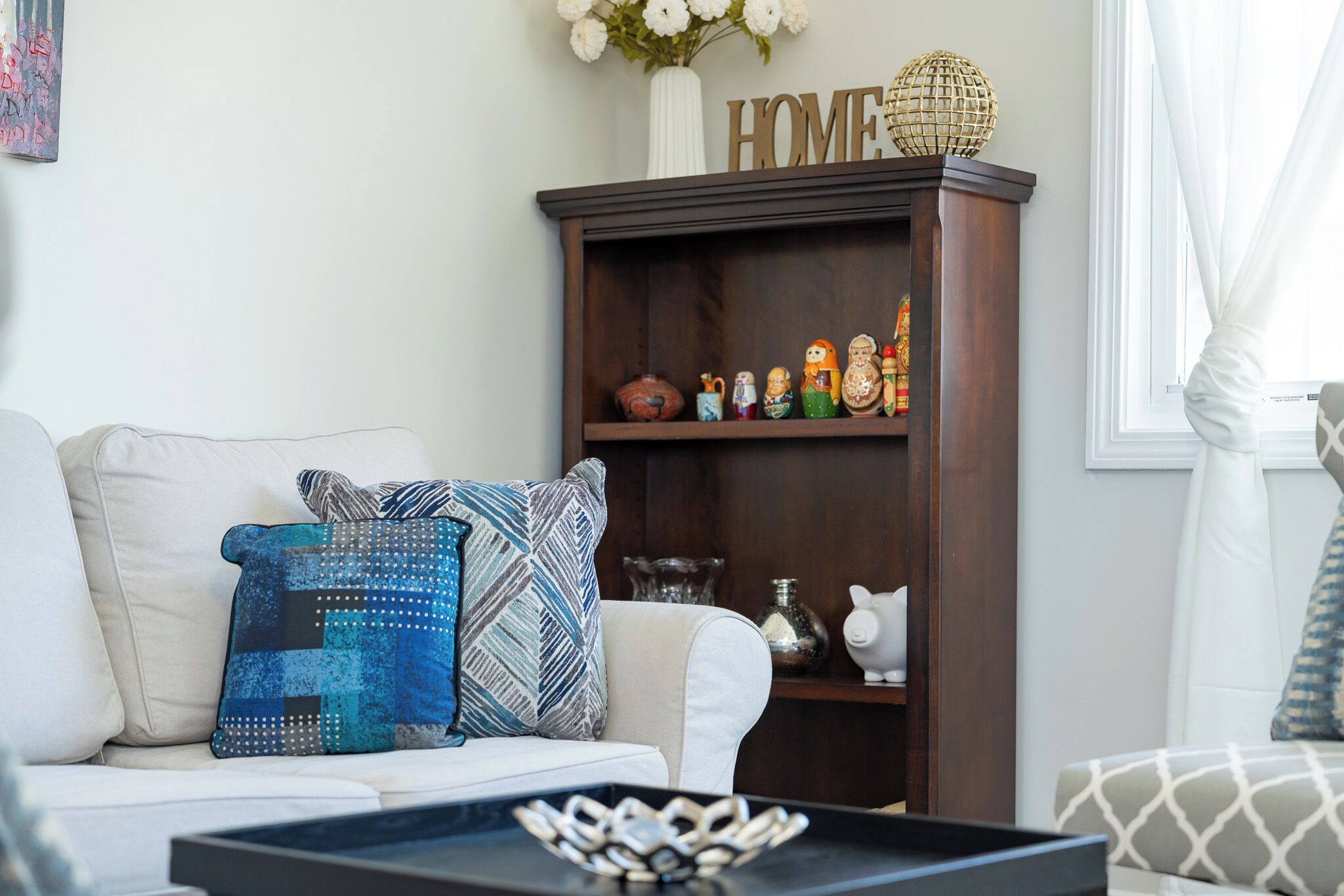 A living room with a white sofa, patterned cushions, a wooden bookshelf with decorative items, and a window with curtains.
