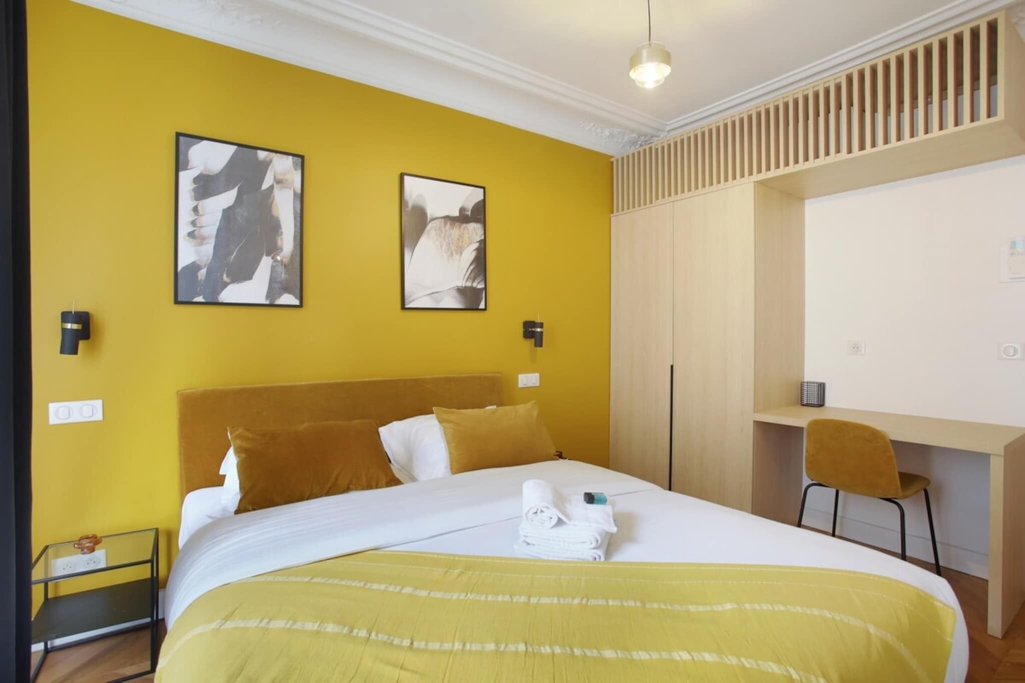 A hotel room with a bed, a desk, and two artwork pieces on the wall.