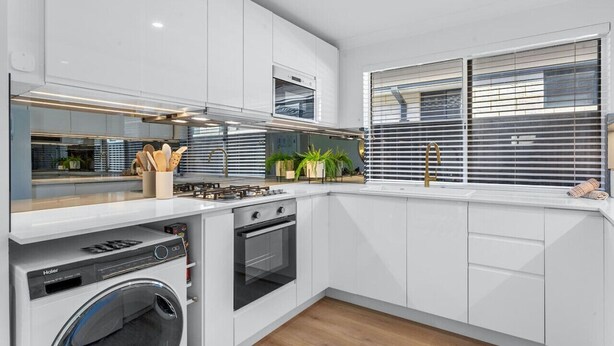 A modern kitchen with white cabinetry, a built-in washer, an oven, and a window with blinds. - Charming Forrestfield Escape! (Forrestfield)