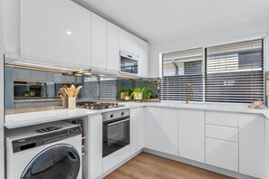 A modern kitchen with white cabinetry, a built-in washer, an oven, and a window with blinds. - Charming Forrestfield Escape! (Forrestfield)