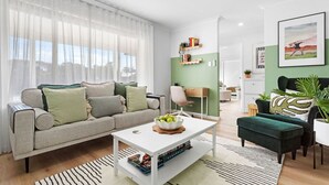 A modern living room with a sofa, coffee table, and green ottoman. There is a bookshelf with books and decorative items, and a painting on the wall. - Charming Forrestfield Escape! (Forrestfield)