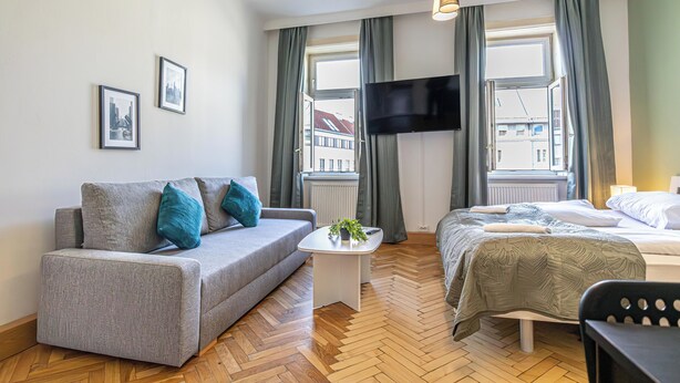 A modern hotel room with a sofa, a bed, a small table, and a TV mounted on the wall. - Comfy 2BR Apt Long Stay Matzleinsdorfer (Vienna)