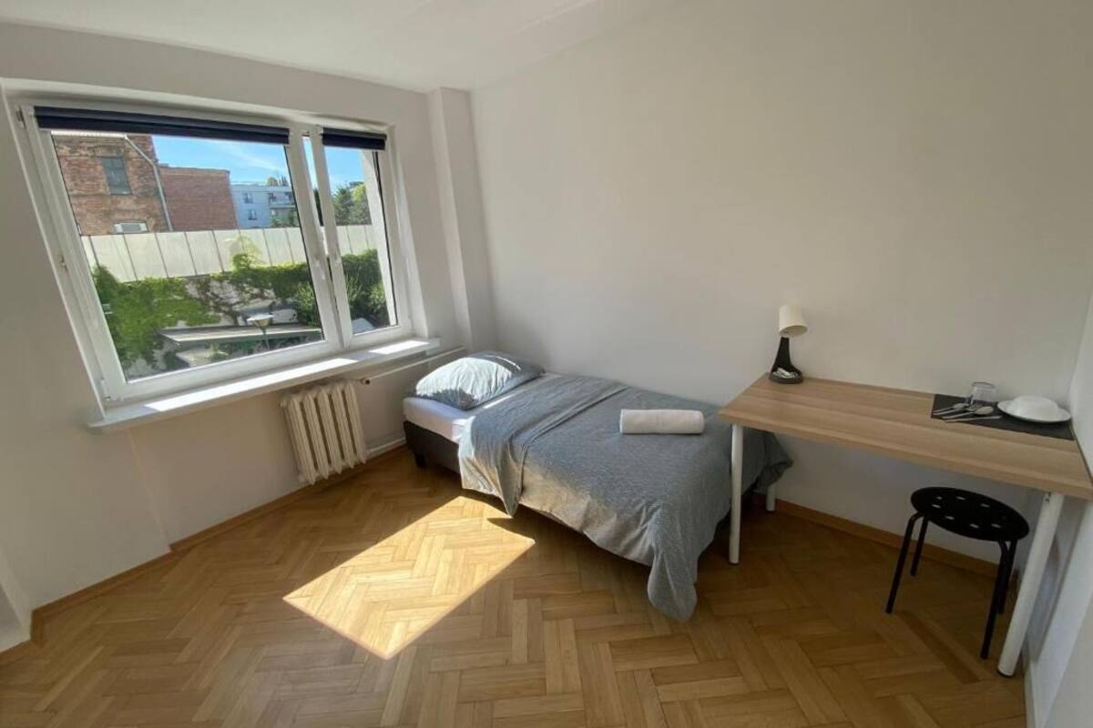 Une petite pièce propre et bien rangée, avec un lit, un bureau et une chaise. Une fenêtre offre une vue sur les bâtiments à l’extérieur.