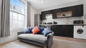 A modern living room with a grey sofa, a washing machine, and a kitchen area with dark cabinets and a marble backsplash. - WhiskAwayHomes Shaw Street Apartment 6 (Worcestershire)