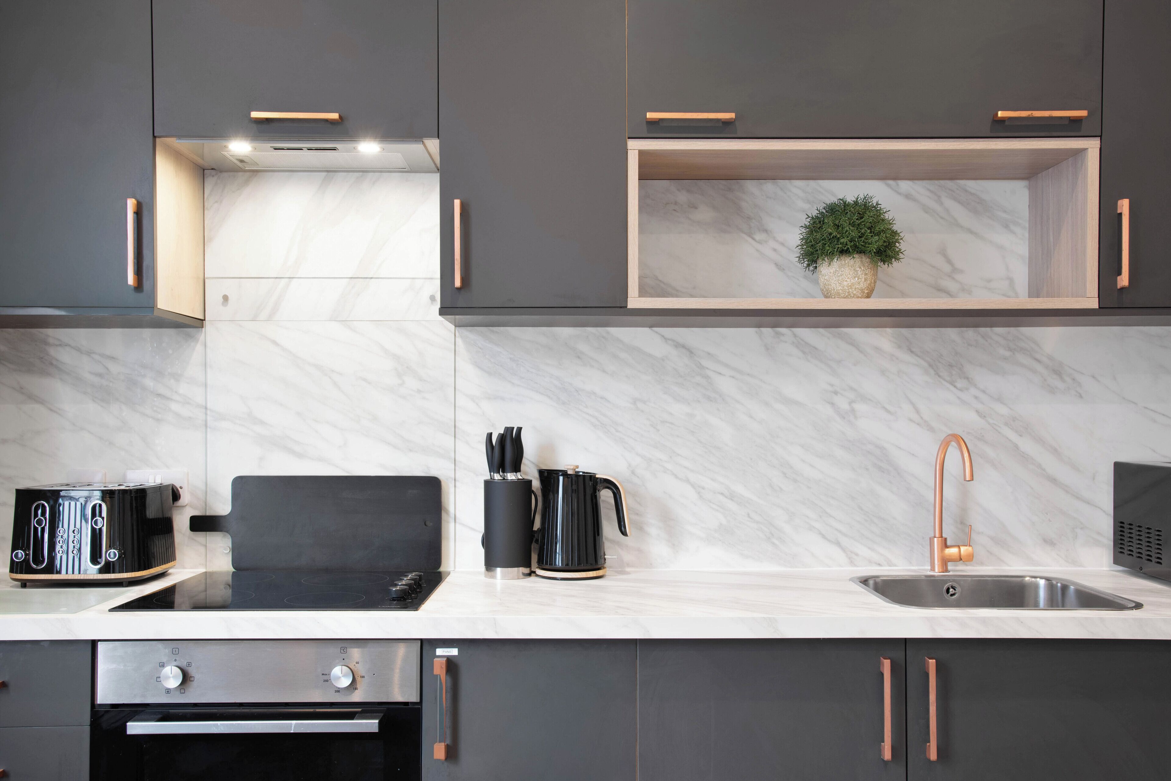 A modern kitchen with dark cabinets, a marble countertop, a built-in oven, a toaster, a coffee maker, and a sink with a copper faucet.