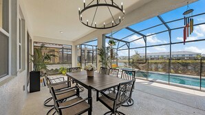 Une vaste terrasse dotée d’une table et de chaises, d’un carillon éolien suspendu, et offrant une vue sur une piscine et un bâtiment en arrière-plan.