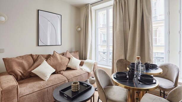 A modern living room with a brown sofa, a round table set with plates and a bottle, and a large window with curtains. - New Apartment Center of Paris Le Marais (Paris)