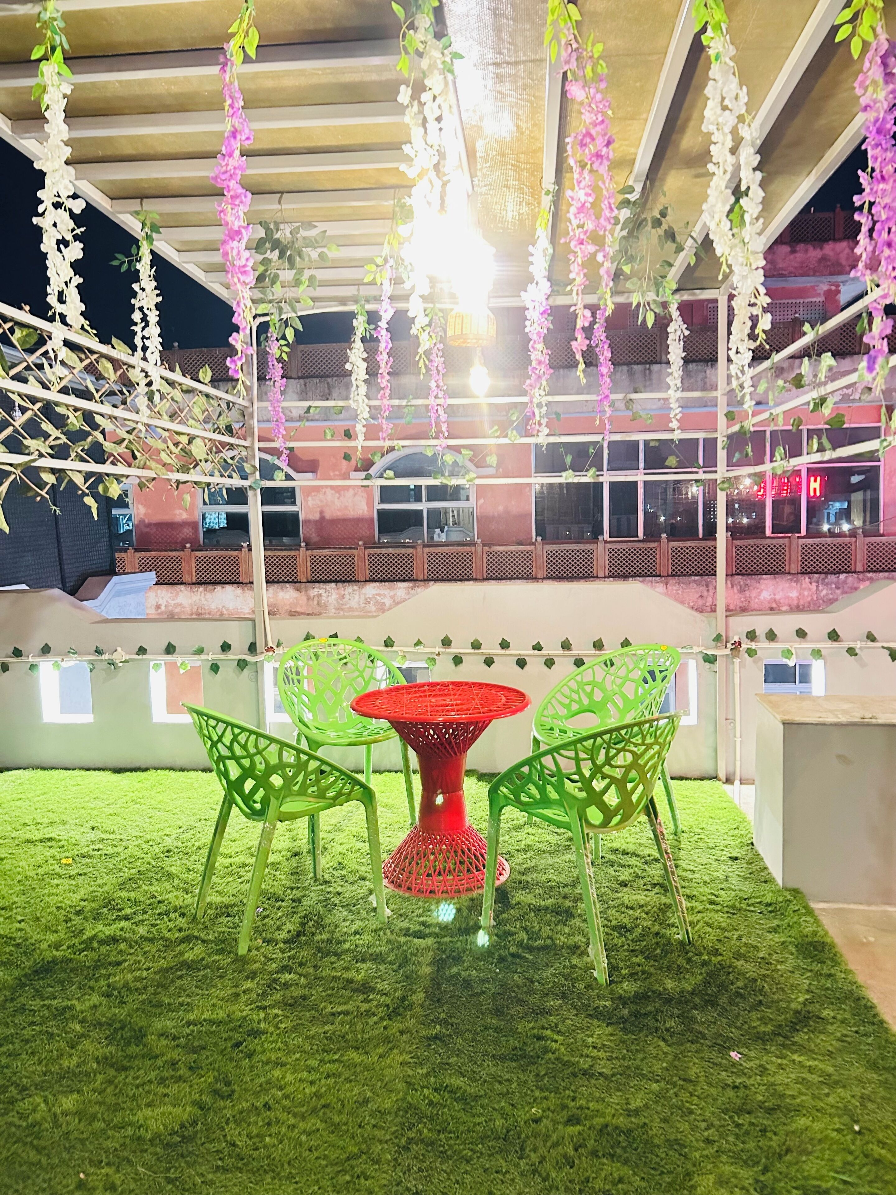 Een terras met groene stoelen en een rode tafel, omgeven door kunstgras en hangende bloemversieringen.