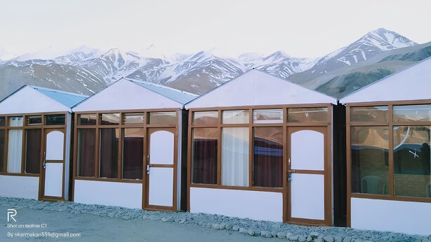 A row of white, wooden cabins with large windows, set against a backdrop of snow-capped mountains. - Pangong View Point Cottage (Leh)