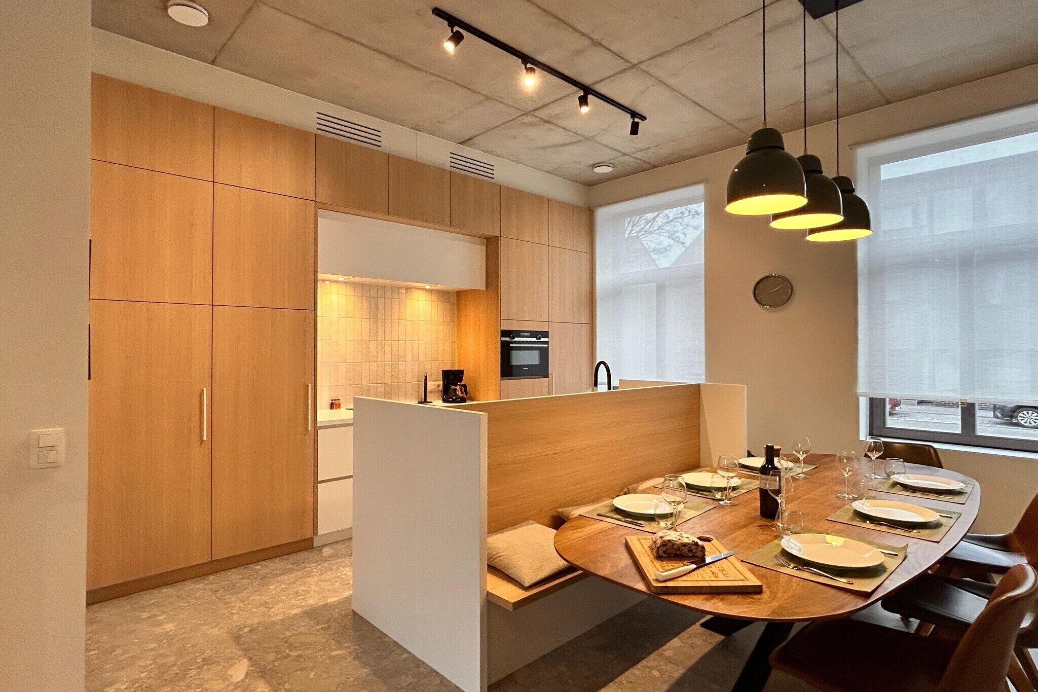Cocina moderna con armarios de madera, comedor con mesa redonda preparada para cuatro personas e iluminación colgante.