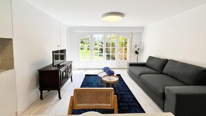 Un salon avec un canapé gris foncé, une table basse en bois, un téléviseur posé sur un meuble en bois foncé et une grande fenêtre donnant sur un paysage verdoyant.