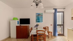 Un coin repas comprenant une table, des chaises, un téléviseur, un réfrigérateur et une fenêtre avec un rideau.