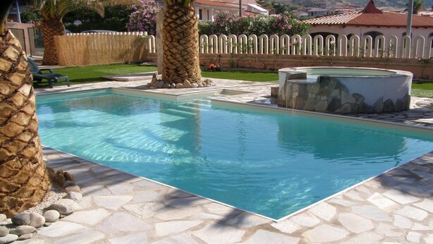A rectangular swimming pool with clear blue water, surrounded by stone tiles and palm trees. - 150 meters from the sea, 2 swimming pools (Valledoria)