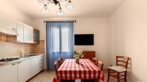 A kitchen with a tiled backsplash, a stove, and a sink. There is a dining table with a checkered tablecloth and chairs. A flat-screen TV is mounted on the wall above the table. - 150 meters from the sea, 2 swimming pools (Valledoria)