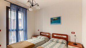 Een kamer met twee bedden, een nachtkastje met een lamp, een raam met blauwe gordijnen en een aan de muur hangend schilderij.