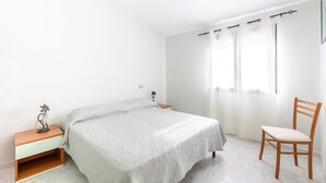 A bedroom with a bed, a nightstand, a chair, and a window with curtains. - 150 meters from the sea, 2 swimming pools (Valledoria)