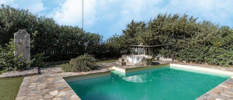 En swimmingpool med stenplattor, omgiven av grönska och en stenkonstruktion.