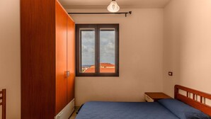 Quarto com cama, guarda-roupa de madeira e janela com vista para edifícios.