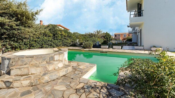 Piscina com degraus de pedra, cercada por vegetação e um edifício branco com varanda.