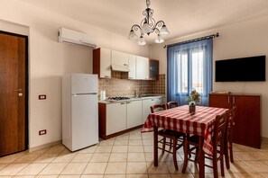 Eine kompakte Küche mit Kühlschrank, Herd und gefliestem Fliesenspiegel. Ein Esstisch mit Stühlen und ein an der Wand befestigtes Fernsehgerät.