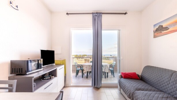A modern living room with a grey sofa, a flat-screen TV, a small table with chairs, and a balcony with a view. - Apartment in with swimming pool and sea view (Castelsardo)