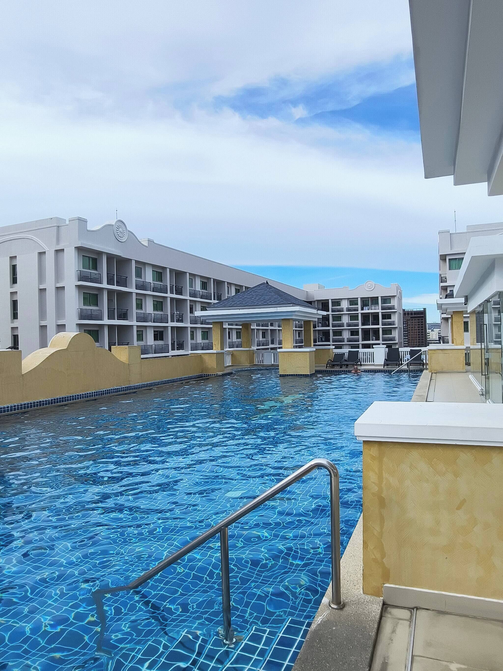 En swimmingpool med en klar, blå vandoverflade, omgivet af moderne bygninger med altaner og et overdækket område.