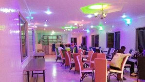 A room with multiple tables and chairs, a ceiling with hanging lights, and a window with blinds. - The Black Gold Hotel Lokichar (Lokichar)
