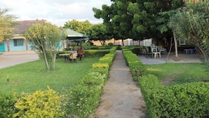 A garden pathway leading to a seating area with tables and chairs, surrounded by greenery and a building with a green roof. - The Black Gold Hotel Lokichar (Lokichar)