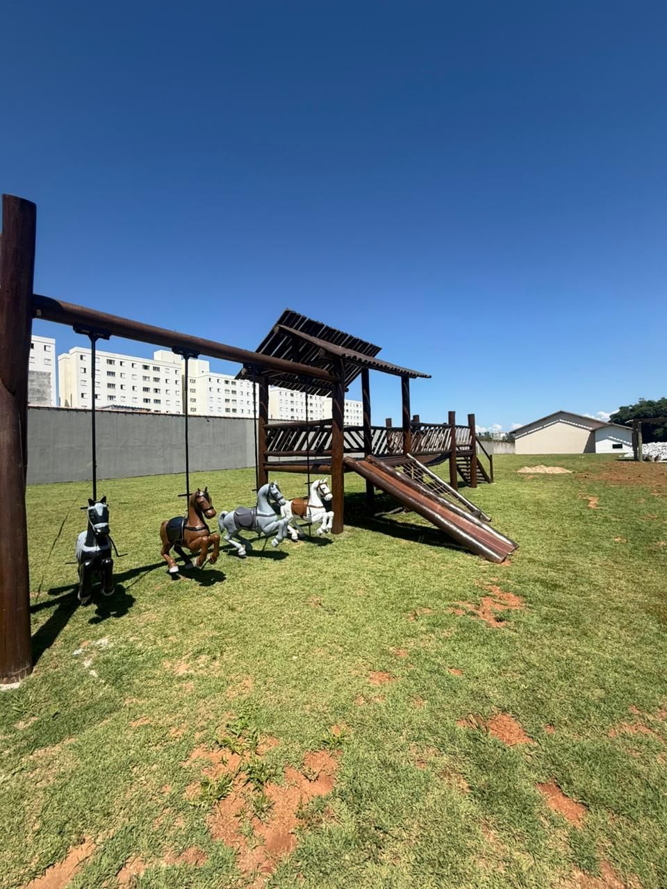 A playground with a wooden structure, including a slide and swings, set against a backdrop of buildings.