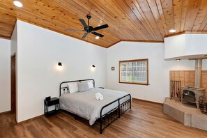 Een slaapkamer met een bed, een plafondventilator, een raam en een houtkachel.