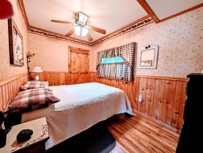 Ein Schlafzimmer mit einem Bett, einem Deckenventilator, einem Fenster mit Karottenvorhängen und Holzwänden.