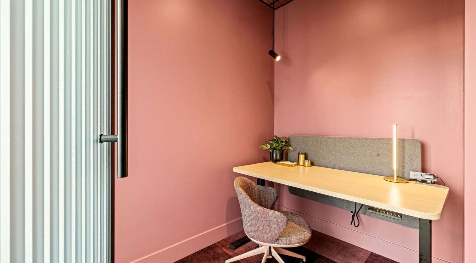 Una habitación pequeña con paredes rosas, una mesa, una silla y una mesita de noche con lámpara.