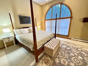 Ein Schlafzimmer mit Himmelbett, Nachttisch, einer Bank, einem großen Fenster mit Jalousien und Teppichboden.