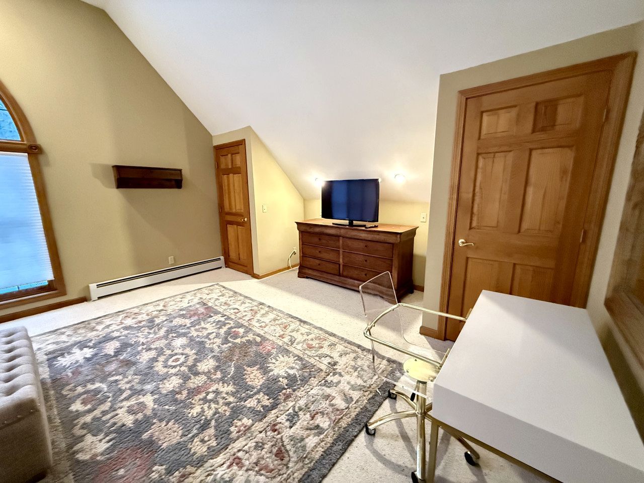 A room with a large window, a wooden door, a dresser, a television on top, and a patterned rug.