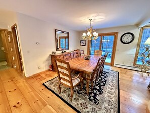 A dining area with a wooden table, chairs, a chandelier, a clock, and a view through a window.