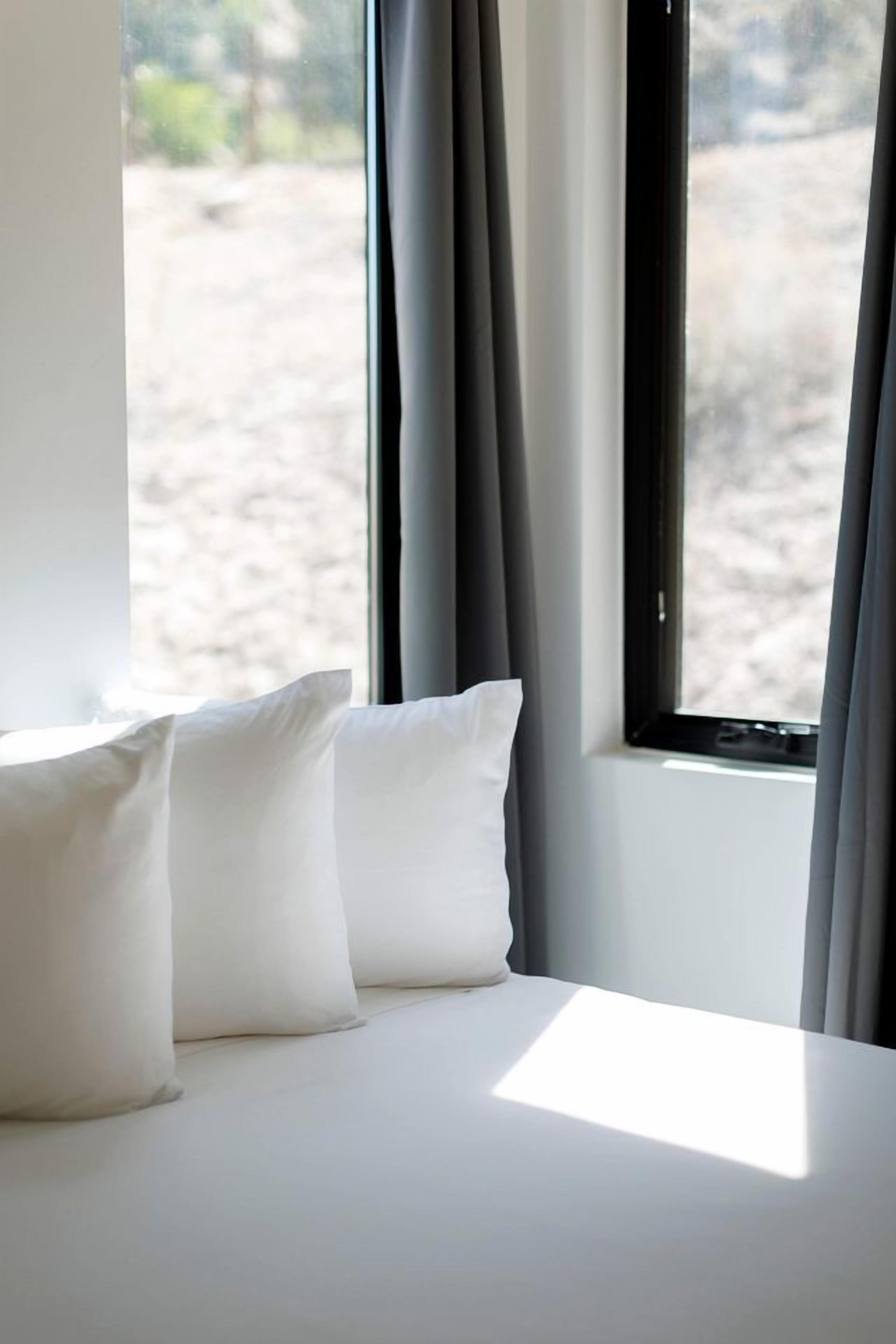 Un lit avec une literie et des oreillers blancs, une fenêtre avec des rideaux et une vue sur un espace extérieur sablonneux.