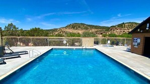 A rectangular swimming pool with clear blue water, surrounded by a fence, and a building with a brick exterior to the right. - Unique Tiny House with Tennis Court in Orderville, Utah (Orderville)