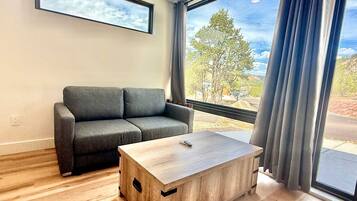 Un salon moderne avec un canapé gris, une table basse en bois et de grandes fenêtres ornées de rideaux.